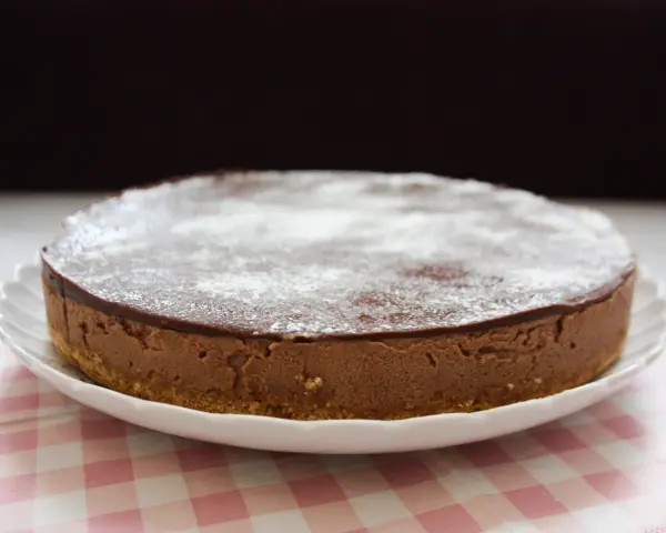 En krämig nutella cheesecake med chokladglasyr och pudersocker, serverad på en vit tallrik med en rosa rutig duk.