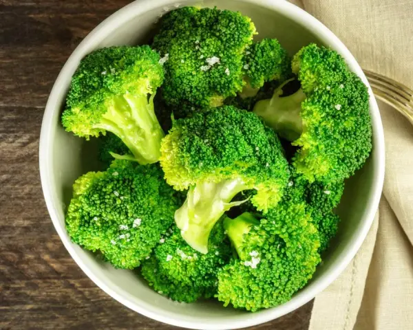 En skål med nykokt, grön broccoli, strödd med flingsalt. En gaffel ligger bredvid på en beige duk.
