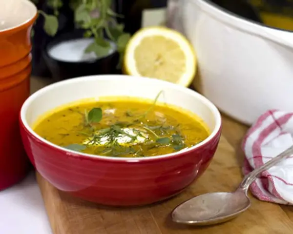 Varm morotssoppa med en klick creme fraiche, garnerad med örter. Serveras i en röd skål med en sked och en citronskiva i bakgrunden.