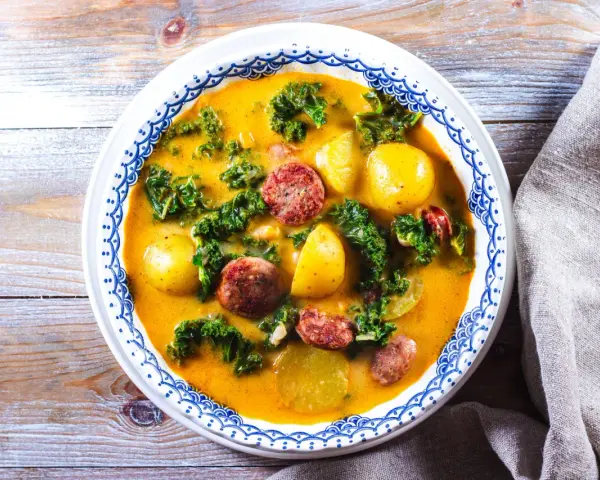 En mustig grönsakssoppa med potatis, grönkål och korvskivor i en gul buljong.