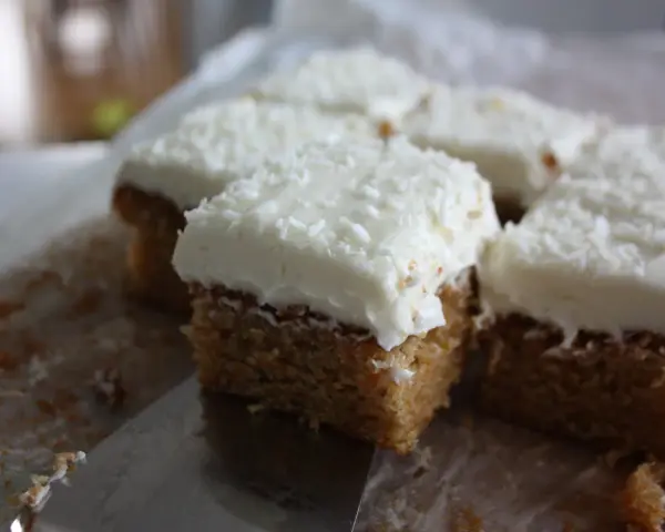 Färska bitar av morotskaka med krämig frosting och kokosflingor. En bit är uppskuren med en kniv.