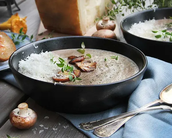 Creamy mushroom soup in a black bowl, topped with grated cheese, mushrooms, and herbs.