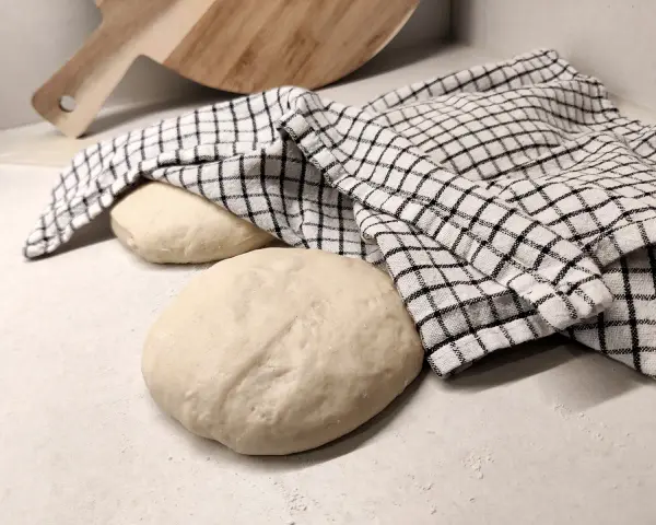 Two balls of pizza dough on a counter, one covered with a checkered cloth, with a wooden pizza peel in the background.