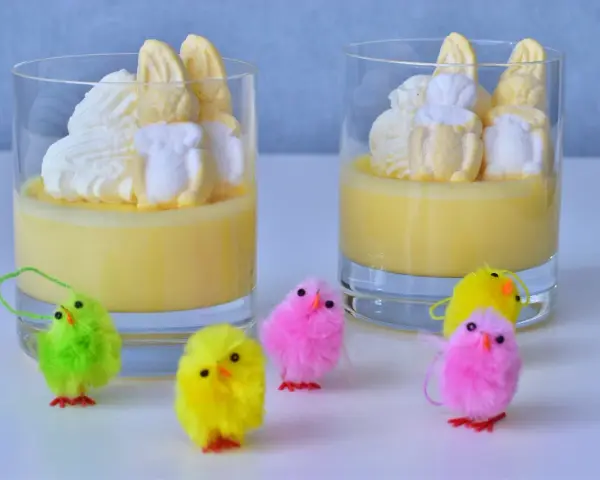 Två glas med gul pannacotta, toppade med vispgrädde och marshmallows som liknar påskkycklingar. Små, fluffiga påskkycklingar i olika färger står framför.