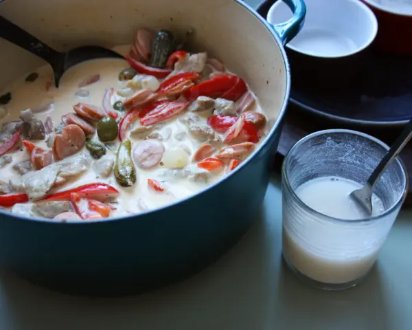En krämig kreolsk gryta med korv, paprika, oliver och pickles i en blå gryta. En sked ligger i ett glas med mjölk.