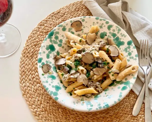 En tallrik med tryffelpasta med svamp och färska örter, serverad på en flätad duk. Ett glas rödvin står bredvid.