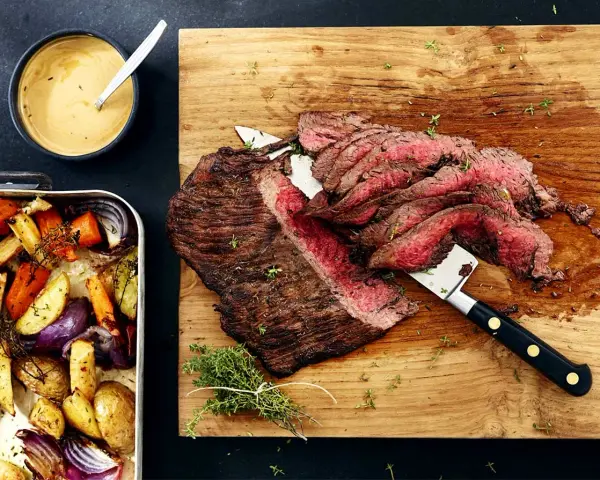 Sliced flank steak with roasted vegetables and a bowl of sauce.