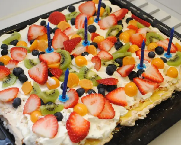 A rectangular meringue cake topped with whipped cream, strawberries, kiwi, blueberries, and physalis, with blue birthday candles.