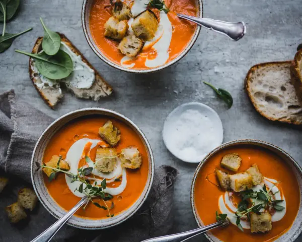 Three bowls of vibrant tomato soup topped with cream, croutons, and herbs, with bread and spinach on the side.