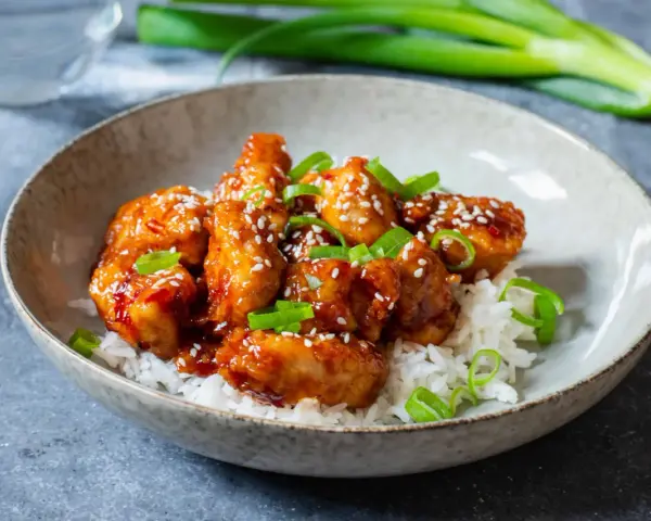 Krispig koreansk kyckling med sesamfrön och salladslök serveras på en bädd av vitt ris i en skål.
