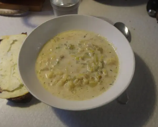 En krämig fänkålssoppa med potatis serveras i en vit skål, bredvid en bit rostat bröd med ost.