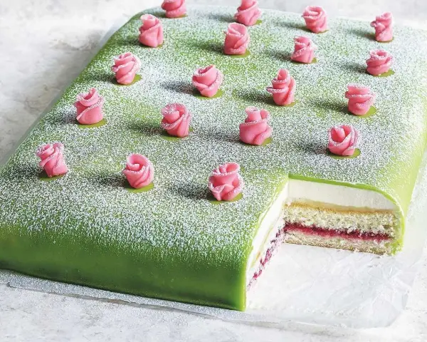 A rectangular green cake topped with powdered sugar and decorated with pink marzipan roses. A slice is cut out, revealing layers of cake, cream, and jam.