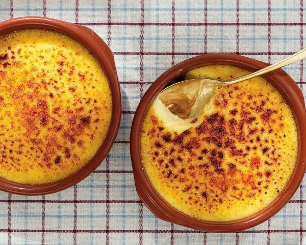 Två krämiga creme brulee i bruna skålar, en med en gyllene sked som lyfter fram den karamelliserade ytan.