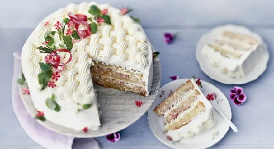 En vacker rabarberkompott tårta med flätmönster av grädde, garnerad med örter och blommor. En bit är upplagd på en tallrik.