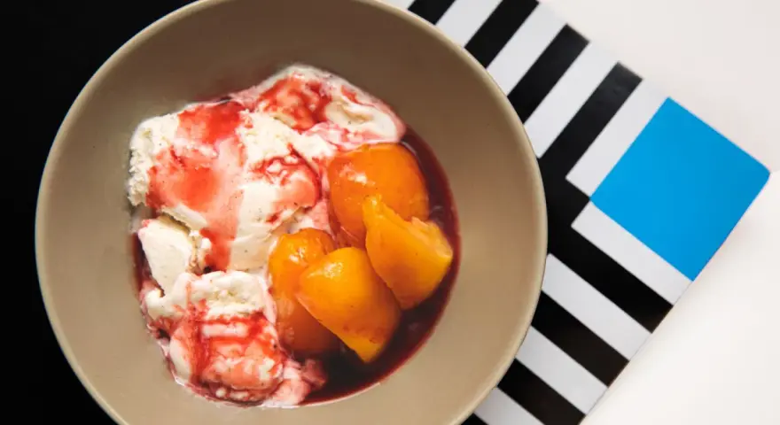 En skål med krämig vaniljglass, saftiga persikohalvor och en röd bärsås. En klassisk peach melba som ser utsökt ut.