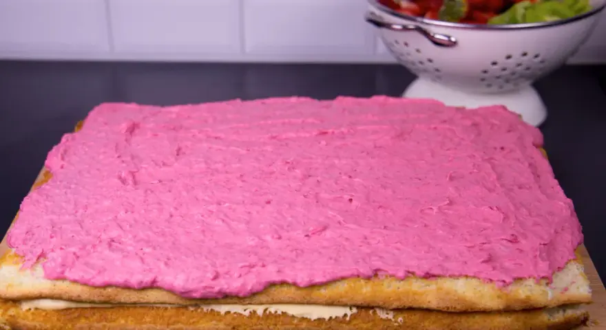 En tårtbotten i långpanna med rosa glasyr. I bakgrunden en skål med jordgubbar och kiwi.