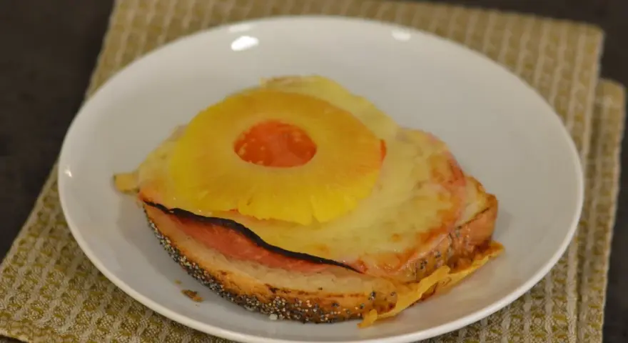 A toasted sandwich with melted cheese, ham, and a pineapple ring on top, served on a white plate.