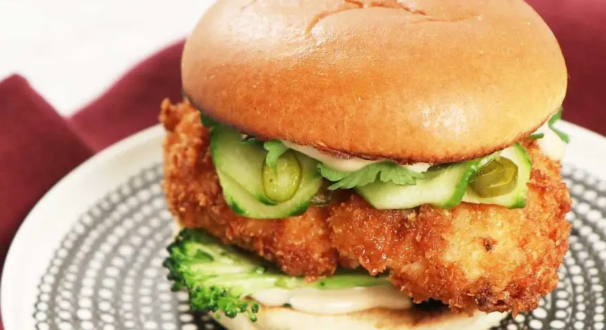 En saftig kyckling hamburgare med krispigt panerat kycklingfilé, gurka, jalapeños och en krämig sås på ett mjukt bröd. Serveras på en snygg tallrik.
