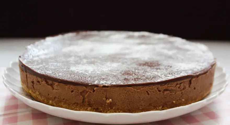 En krämig nutella cheesecake med chokladglasyr och pudersocker, serverad på en vit tallrik med en rosa rutig duk.
