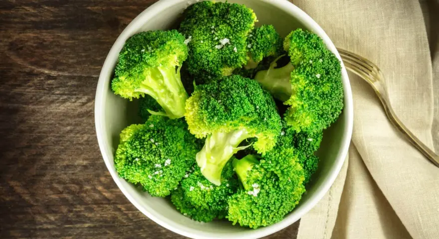 En skål med nykokt, grön broccoli, strödd med flingsalt. En gaffel ligger bredvid på en beige duk.