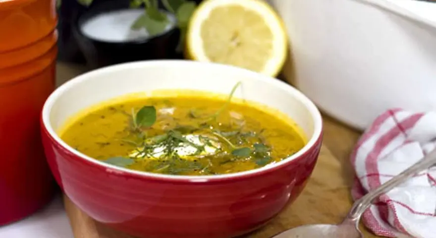 Varm morotssoppa med en klick creme fraiche, garnerad med örter. Serveras i en röd skål med en sked och en citronskiva i bakgrunden.
