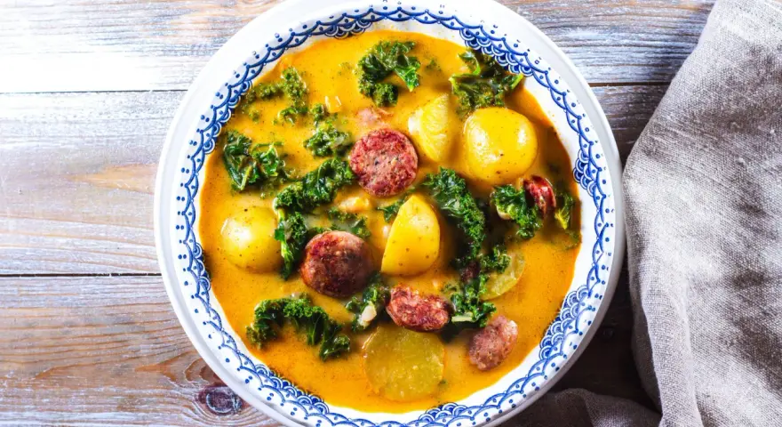 En mustig grönsakssoppa med potatis, grönkål och korvskivor i en gul buljong.