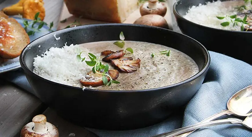 Creamy mushroom soup in a black bowl, topped with grated cheese, mushrooms, and herbs.