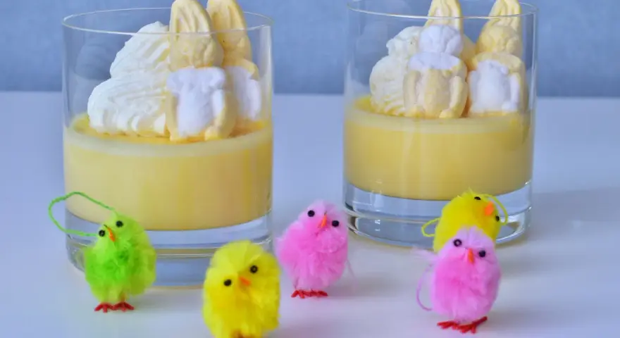 Två glas med gul pannacotta, toppade med vispgrädde och marshmallows som liknar påskkycklingar. Små, fluffiga påskkycklingar i olika färger står framför.