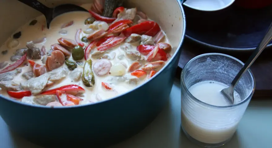 En krämig kreolsk gryta med korv, paprika, oliver och pickles i en blå gryta. En sked ligger i ett glas med mjölk.