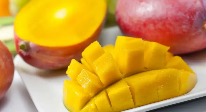 En hel och en halverad mango, skuren i bitar, ligger på en vit tallrik. Bakom syns druvor och en annan hel mango.