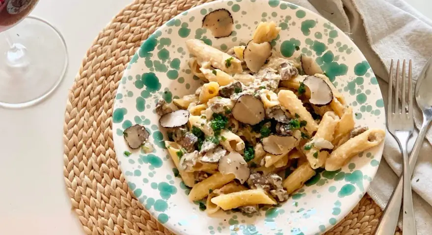 En tallrik med tryffelpasta med svamp och färska örter, serverad på en flätad duk. Ett glas rödvin står bredvid.