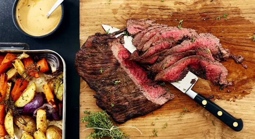 Sliced flank steak with roasted vegetables and a bowl of sauce.