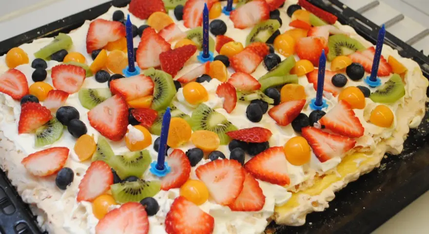 A rectangular meringue cake topped with whipped cream, strawberries, kiwi, blueberries, and physalis, with blue birthday candles.