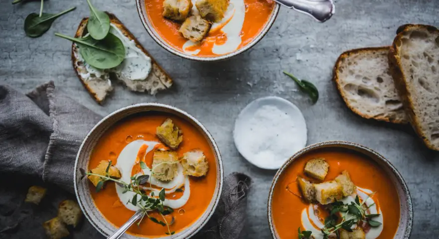 Three bowls of vibrant tomato soup topped with cream, croutons, and herbs, with bread and spinach on the side.