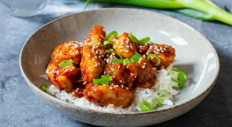 Krispig koreansk kyckling med sesamfrön och salladslök serveras på en bädd av vitt ris i en skål.