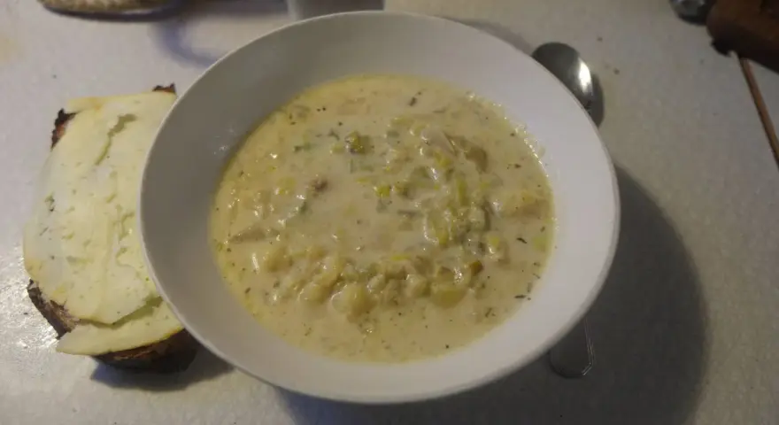 En krämig fänkålssoppa med potatis serveras i en vit skål, bredvid en bit rostat bröd med ost.