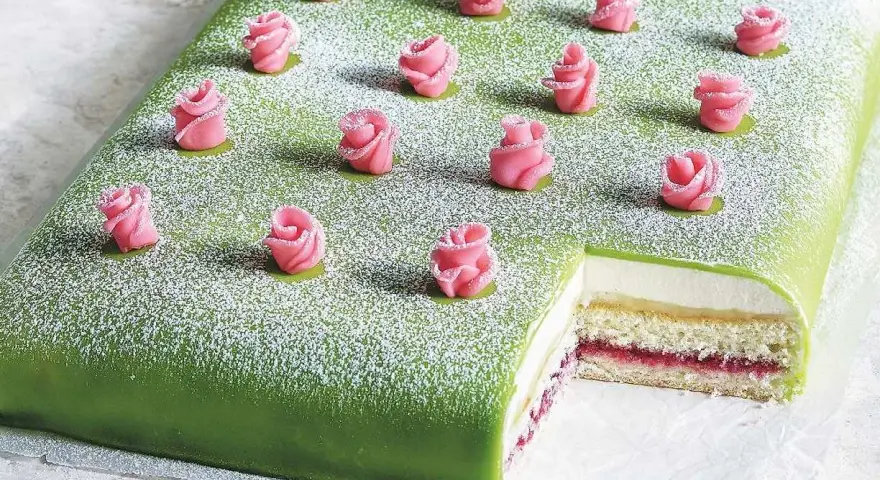 A rectangular green cake topped with powdered sugar and decorated with pink marzipan roses. A slice is cut out, revealing layers of cake, cream, and jam.