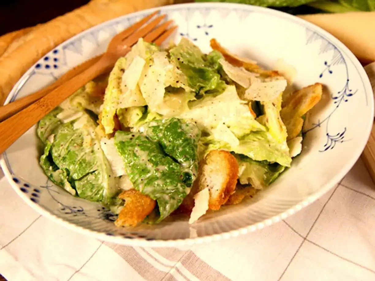 A close-up of a classic Caesar salad with romaine lettuce, croutons, and shaved Parmesan cheese, served in a white bowl with blue trim.