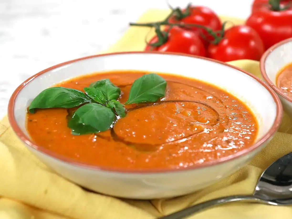 En skål med krämig rostad tomatsoppa, garnerad med färsk basilika och en virvel av olivolja. Bakom syns färska tomater.
