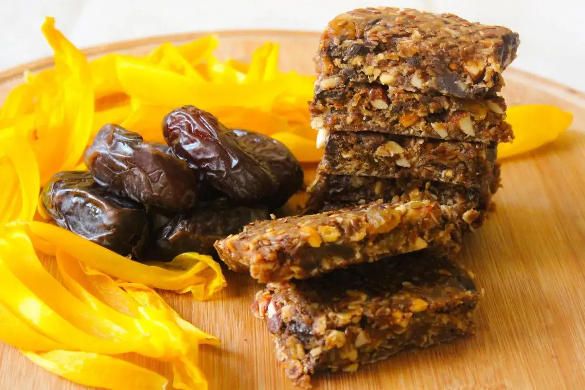En hög med hemgjorda energibars gjorda på dadlar nyttigt, nötter och frön, bredvid färska dadlar och gula mangostrimlor på en träskärbräda.