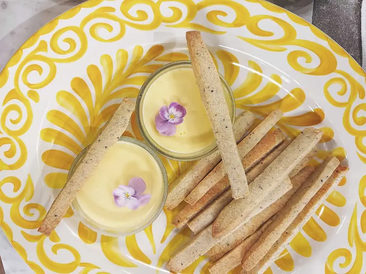 Två små skålar med gul kräm, dekorerade med violer, serveras med krispiga kex på en gulmönstrad tallrik. En perfekt påsk dessert.