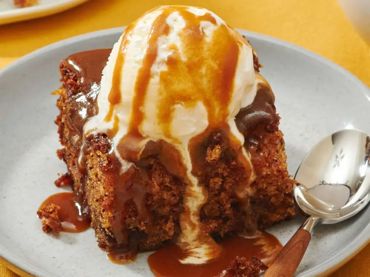 En bit saftig sticky toffee pudding med vaniljglass och kolas&aring;s som rinner ner. En sked ligger bredvid.