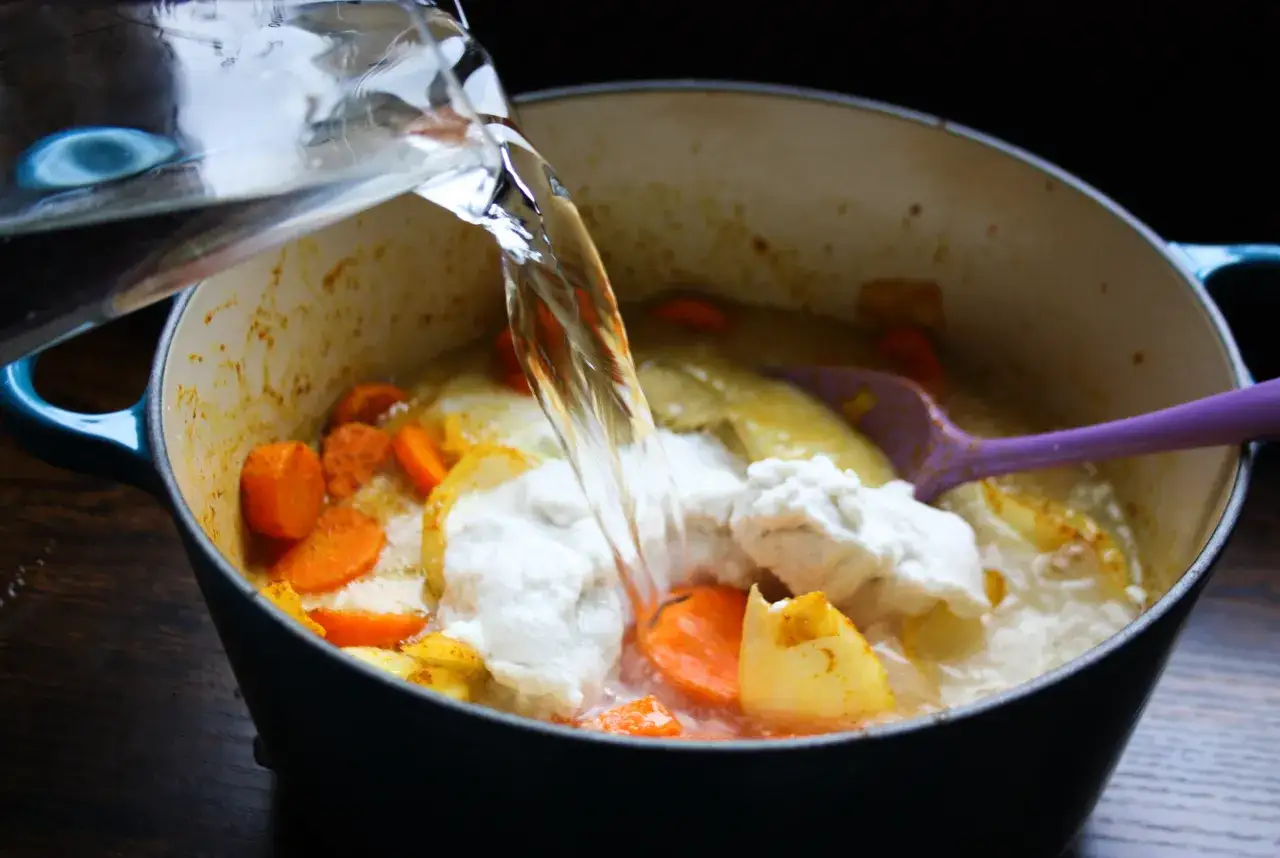 Vatten hälls i en gryta med morötter och potatis för en enkel morotssoppa. En lila slev ligger i grytan.