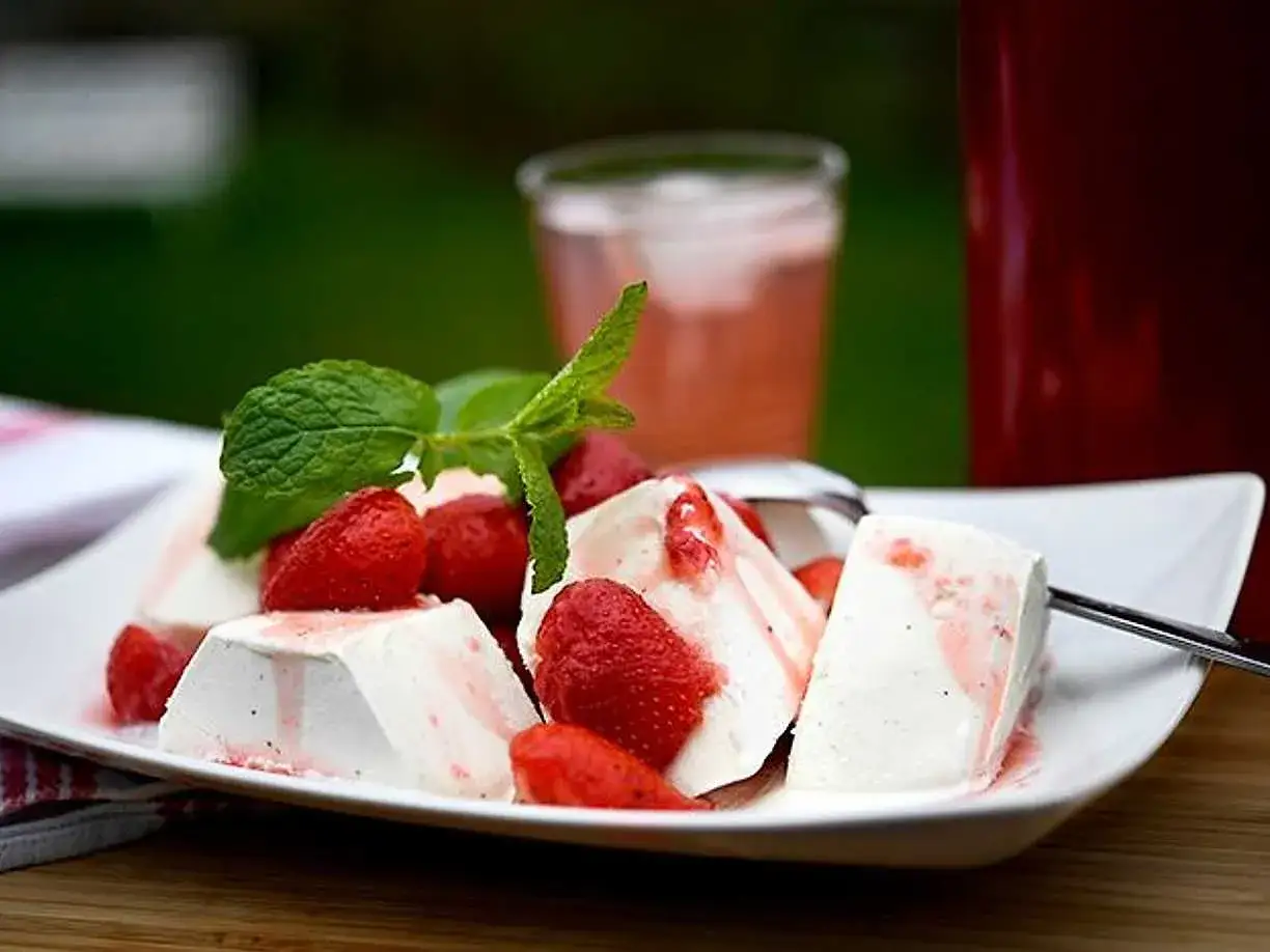 En krämig dessert med marinerade jordgubbar och mynta på en vit tallrik. En glasburk med rosa dryck och en röd mugg i bakgrunden.