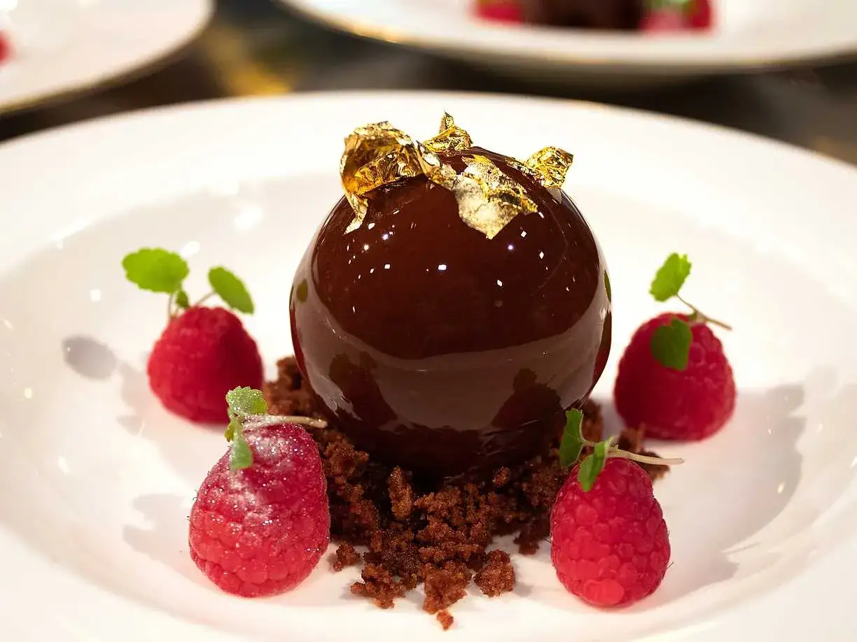 En glänsande chokladboll med guldfolie, omgiven av hallon och chokladsmulor. Perfekt dessert för nyårsafton.