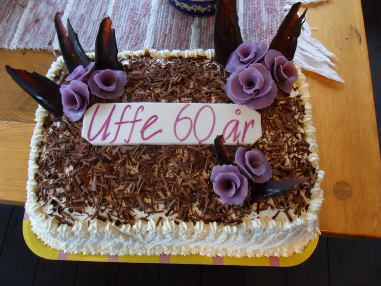 En stor tårtbotten i långpanna, dekorerad med chokladspån, grädde och lila blommor. Grattis till Uffe 60 år!