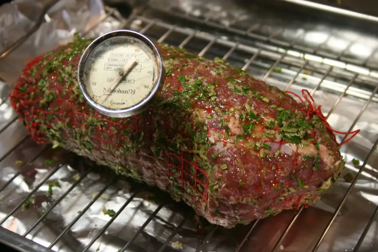 En fransyska i ugn, förberedd för låg temperatur. Köttet är nätat och har en örtmarinad. En köttermometer visar temperaturen.