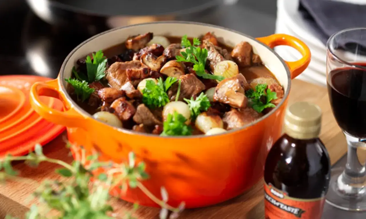 En mustig boeuf bourguignon i orange gryta, med champinjoner, lök och persilja. Ett glas rödvin bredvid.