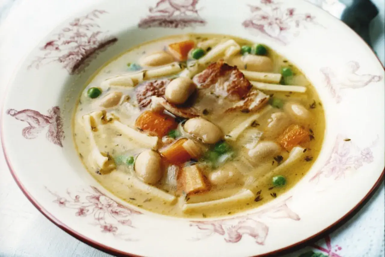 En sk&aring;l med minestronesoppa med fl&auml;sk, b&ouml;nor, mor&ouml;tter, &auml;rtor och pasta. Tallriken har en dekorativ kant med r&ouml;da m&ouml;nster.