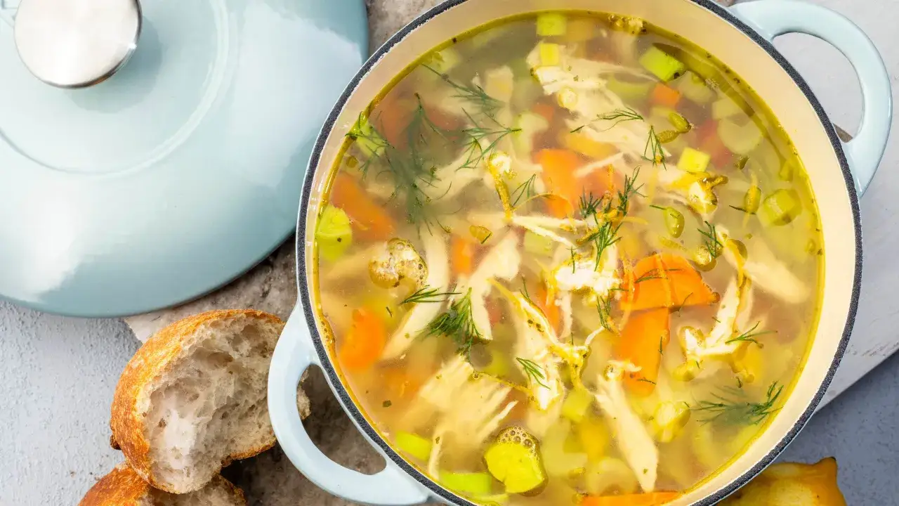 En värmande kycklingsoppa med morötter, selleri och dill i en ljusblå gryta, bredvid ett nybakat bröd.